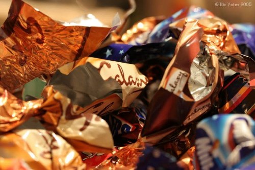 Close-up image showing a pile of crumpled recyclable foil wrappers and chocolate packaging materials, primarily in metallic copper, silver, and vibrant blue tones. The wrappers display creases and folds, with some featuring partially visible branding and text. They are scattered on a flat surface, possibly in a domestic or commercial setting, with a warm, soft lighting that highlights the reflective qualities of the foil finishes. The background is blurred, keeping the focus on the piled packaging, which appears to be ready for disposal or collection by a garden clearance service such as Garden Clearance Turnham Green. This scene illustrates typical waste materials associated with rubbish removal in a garden or household context within London, subtly reinforcing the need for professional rubbish clearance to manage recyclable waste efficiently.
