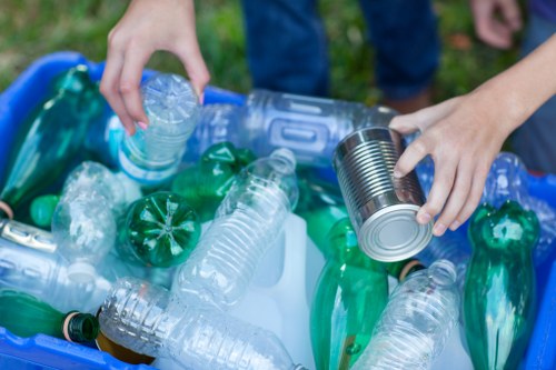 A close-up view of a personâ€™s hands organizing and handling various recyclable plastic bottles and a metal can, placed on top of a blue plastic recycling bin. The bottles are primarily transparent and green, with some caps visible, indicating different types of plastic. The metal can, which appears to be a food or drink container, is held securely in one hand while the other hand reaches towards additional items. The scene is set outdoors, with a grassy background and the individual's jeans visible, suggesting a garden clearance or waste collection activity typical of services offered by Garden Clearance Turnham Green. The lighting is natural, highlighting the textures and colors of the plastic and metallic objects, emphasizing the focus on waste separation and collection for rubbish removal in the local area. The image illustrates the sorting process involved in environmentally conscious rubbish disposal or recycling efforts.
